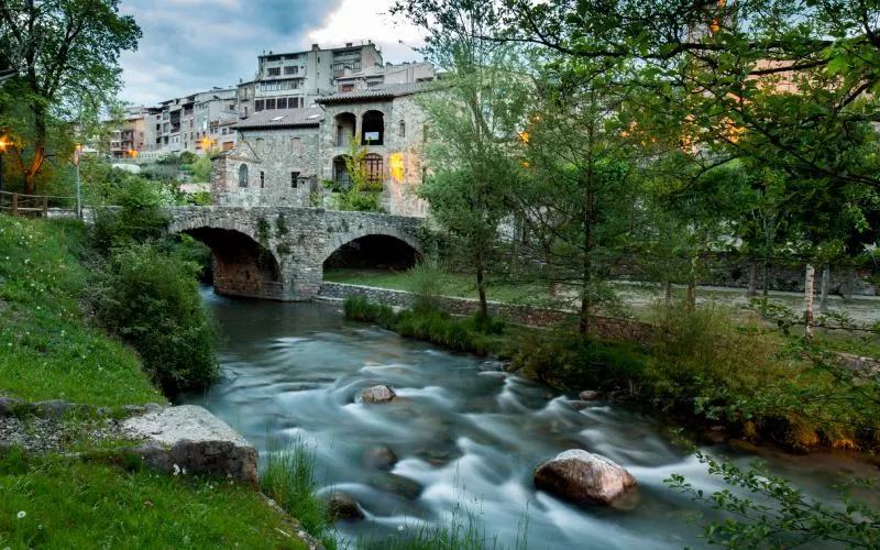 A river surrounded by grass and trees. In the background, a bridge and some houses. 